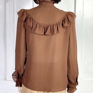 Puede incluir: Una blusa marr&oacute;n de gasa con un cuello alto con volantes y pu&ntilde;os con volantes. La blusa tiene un gran volante en la espalda.