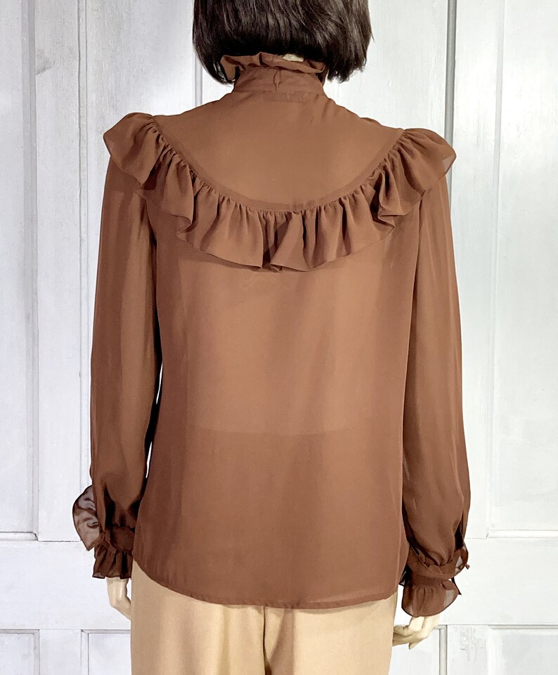 Puede incluir: Una blusa marr&oacute;n de gasa con un cuello alto con volantes y pu&ntilde;os con volantes. La blusa tiene un gran volante en la espalda.