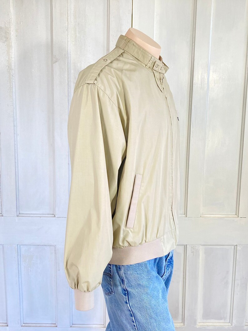May include: A beige lightweight jacket with a zipper closure and a stand-up collar. The jacket has a single pocket on the left side and a ribbed waistband.