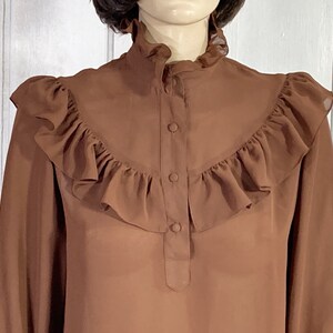 Puede incluir: Una blusa marr&oacute;n de gasa con un cuello alto con volantes y cierre de botones en la parte delantera. La blusa tiene volantes a lo largo del escote y los hombros.
