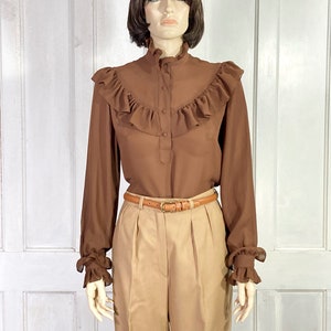 Puede incluir: Una blusa marr&oacute;n de manga larga con cuello alto con volantes y pu&ntilde;os con volantes. La blusa tiene cierre de botones en la parte delantera y un cintur&oacute;n marr&oacute;n.