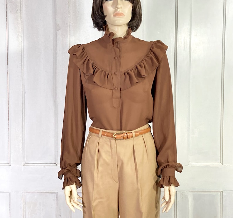 Puede incluir: Una blusa marr&oacute;n de manga larga con cuello alto con volantes y pu&ntilde;os con volantes. La blusa tiene cierre de botones en la parte delantera y un cintur&oacute;n marr&oacute;n.
