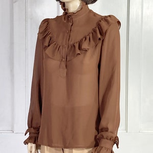 Puede incluir: Una blusa marr&oacute;n de gasa con cuello alto con volantes y mangas largas. La blusa tiene un cierre de botones en la parte delantera y pu&ntilde;os con volantes.