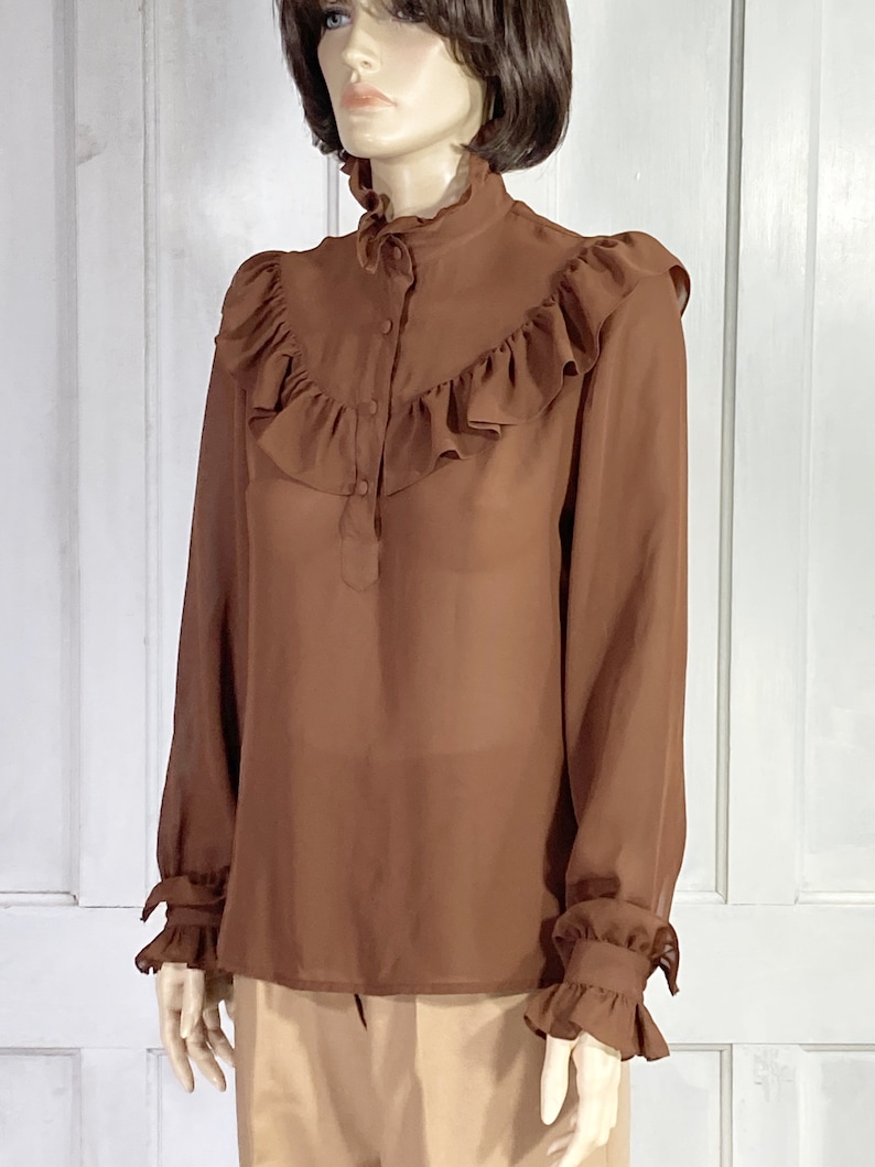 Puede incluir: Una blusa marr&oacute;n de gasa con cuello alto con volantes y mangas largas. La blusa tiene un cierre de botones en la parte delantera y pu&ntilde;os con volantes.