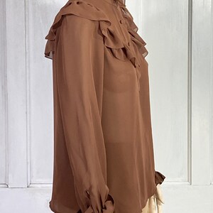 Puede incluir: Una blusa marr&oacute;n de manga larga con un cuello alto con volantes y pu&ntilde;os con volantes. La blusa est&aacute; hecha de una tela transparente.