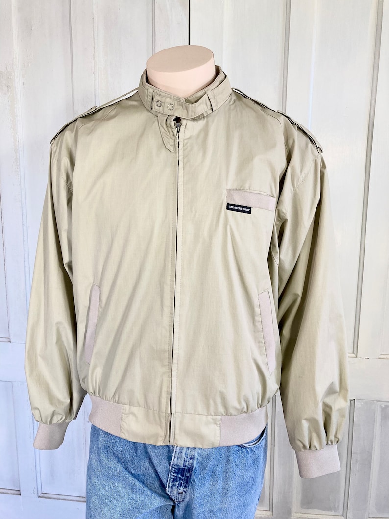May include: A beige Members Only jacket with a zipper closure and a ribbed waistband. The jacket has a stand-up collar and two pockets on the front.