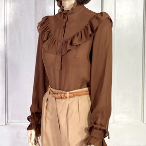 Puede incluir: Una blusa marr&oacute;n de manga larga con cuello alto con volantes y pu&ntilde;os con volantes. La blusa est&aacute; hecha de una tela transparente y tiene un cierre de botones en la parte delantera.