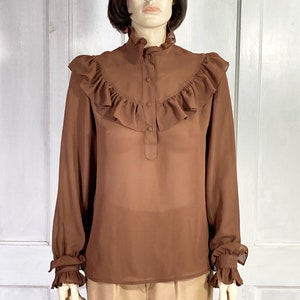Puede incluir: Una blusa marr&oacute;n de gasa con un cuello alto con volantes y mangas largas. Las mangas tienen volantes en los pu&ntilde;os. La blusa tiene un cierre de botones en la parte delantera.