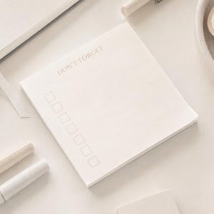 May include: A square off-white notepad featuring the text DON'T FORGET printed in a thin font at the top, followed by a vertical column of seven empty square checkboxes for list-making, presented on a neutral-toned surface.