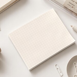 May include: A rectangular notepad with a cream-colored grid pattern on white paper. The notepad is surrounded by stationery items including a Milk Tea highlighter, a roll of plaid washi tape, and a small bowl of white paper clips.