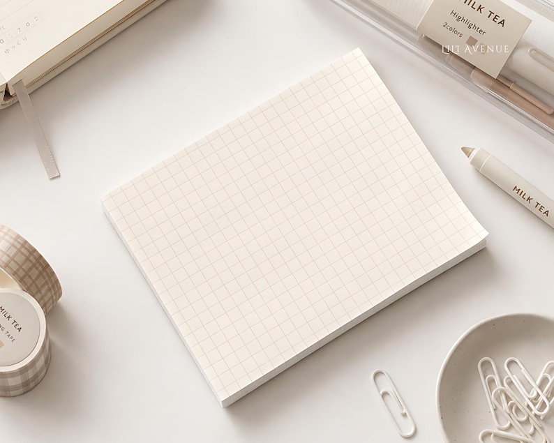 May include: A rectangular notepad with a cream-colored grid pattern on white paper. The notepad is surrounded by stationery items including a Milk Tea highlighter, a roll of plaid washi tape, and a small bowl of white paper clips.