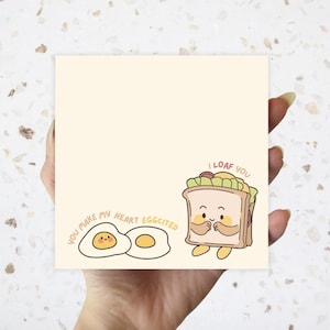 May include: A square, cream-colored notepad with cute food illustrations. One side features a smiling sandwich with the text "I loaf you." The other side has two fried eggs and the text "You make my heart eggcited."