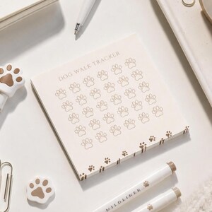 May include: A square notepad with a cream-colored background featuring the text DOG WALK TRACKER at the top. The page contains a grid of thirty-five brown outlined paw prints, with additional small paw print icons along the bottom edge.