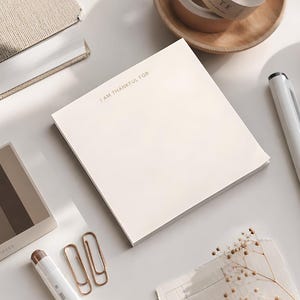May include: A square notepad with cream-colored paper featuring the text I AM THANKFUL FOR printed in a small, dark font at the top. The notepad is positioned among various stationery items including pens, paper clips, and a small color swatch card.
