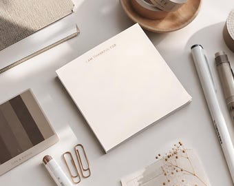 May include: A square notepad with cream-colored paper featuring the text I AM THANKFUL FOR printed in a small, dark font at the top. The notepad is positioned among various stationery items including pens, paper clips, and a small color swatch card.