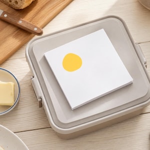 May include: A square, light beige plastic container with a flat lid, topped with a stack of white square paper notes featuring a single, solid yellow circle graphic printed on the top sheet, positioned centrally on the lid surface.