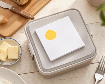 May include: A square, light beige plastic container with a flat lid, topped with a stack of white square paper notes featuring a single, solid yellow circle graphic printed on the top sheet, positioned centrally on the lid surface.