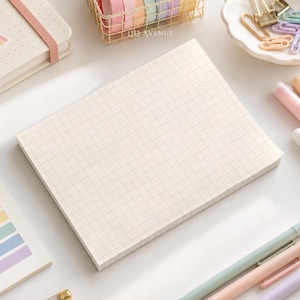 May include: A rectangular notepad with a light cream grid pattern, surrounded by stationery items including a rainbow-themed notebook, pastel pens, a wire basket of washi tape, a heart-shaped paperclip, and a dish of colorful paperclips.