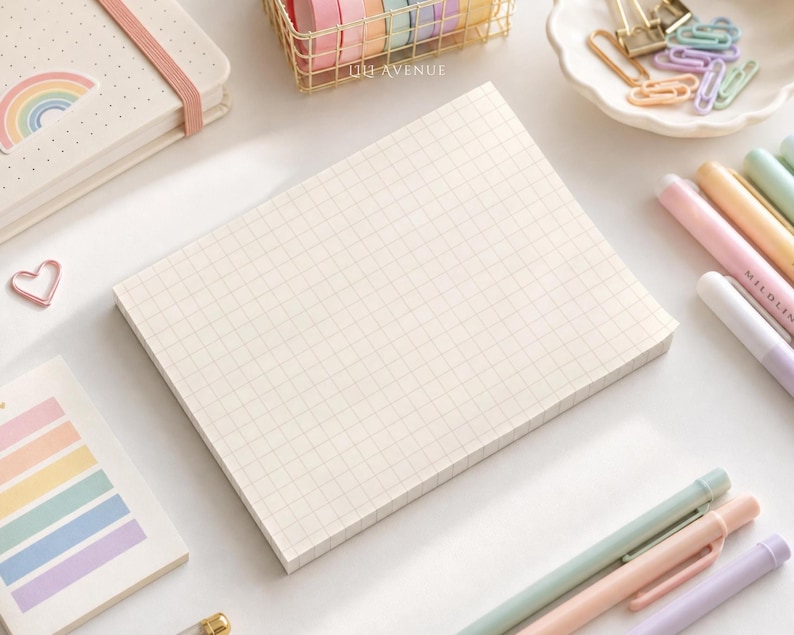 May include: A rectangular notepad with a light cream grid pattern, surrounded by stationery items including a rainbow-themed notebook, pastel pens, a wire basket of washi tape, a heart-shaped paperclip, and a dish of colorful paperclips.