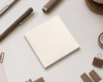 May include: A square notepad with cream-colored paper featuring thin horizontal lines. The notepad is surrounded by various stationery items including brown pens, a brown binder clip, and a small portion of a spiral-bound notebook.