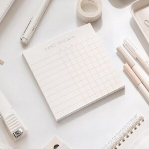 May include: A square habit tracker notepad with a grid layout and the text HABIT TRACKER printed at the top. The notepad features light gray lines on a white background, designed for daily tracking of tasks or habits in a clean, organized format.