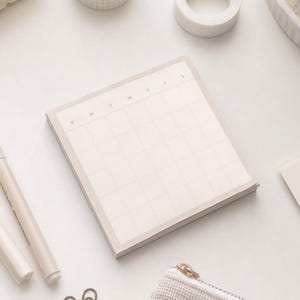 May include: A square notepad with a light beige grid pattern and a calendar layout. The top row features the letters S, M, T, W, T, F, S printed in a minimalist font. The paper is off-white with a subtle border, designed for planning and scheduling.