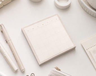 May include: A square notepad with a light beige grid pattern and a calendar layout. The top row features the letters S, M, T, W, T, F, S printed in a minimalist font. The paper is off-white with a subtle border, designed for planning and scheduling.