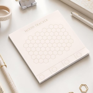 May include: A square paper notepad with a light cream background featuring the text MOOD TRACKER at the top. The center contains a grid of thin-lined hexagonal shapes arranged in a honeycomb pattern, with additional individual hexagons at the bottom.