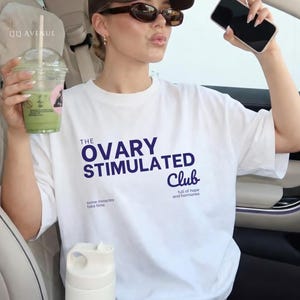 Op de afbeelding: Wit T-shirt met marineblauwe tekst "THE OVARY STIMULATED Club". Het shirt heeft ook de zinnen "some miracles take time" en "full of hope and hormones". Een persoon houdt een groene drank en een telefoon vast.