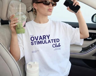 May include: White t-shirt with the text "THE OVARY STIMULATED Club" in navy blue. The shirt also has the phrases "some miracles take time" and "full of hope and hormones". The person is holding a green drink and a phone.