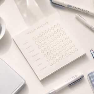 May include: A square notepad with the text WATER TRACKER printed at the top, featuring seven rows labeled with days of the week from Sunday to Saturday, each containing eight outlined water drop icons for tracking daily water consumption.
