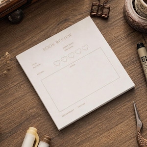 May include: A square white paper notepad featuring a printed book review template. The text includes BOOK REVIEW, START DATE, END DATE, TITLE, AUTHOR, RATING, NOTES, and RECOMMEND YES/NO. Five heart outlines are positioned below the header.