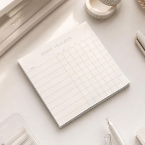 May include: A square paper habit tracker notepad with a cream-colored background and thin gray grid lines. The top of the page features the text HABIT TRACKER in a simple font, with columns marked for days of the week and rows for daily tasks.