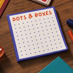K&ouml;nnte beinhalten: Ein quadratischer Notizblock mit wei&szlig;em Hintergrund und blauem Rand, der oben den orangefarbenen Text DOTS & BOXES und ein auf die Seite gedrucktes Raster aus blauen Punkten aufweist. Der Notizblock liegt auf einer Holzoberfl&auml;che mit Spielsteinen.