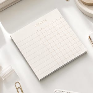May include: A square notepad with a cream-colored paper surface featuring a grid layout. The top section is printed with the word TRACKER in capital letters, followed by the letters S, M, T, W, T, F, S representing the days of the week.