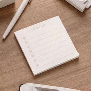 May include: A square off-white notepad with the text TO DO LIST printed at the top. The notepad features six horizontal lines, each preceded by a small light gray heart icon, designed for organizing tasks and daily notes on a flat surface.