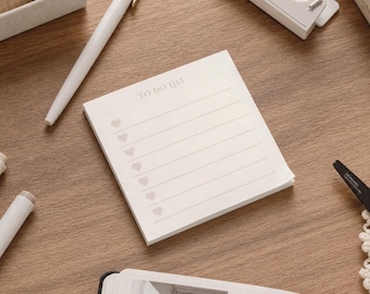 May include: A square off-white notepad with the text TO DO LIST printed at the top. The notepad features six horizontal lines, each preceded by a small light gray heart icon, designed for organizing tasks and daily notes on a flat surface.