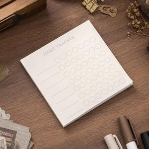 May include: A square white paper habit tracker notepad featuring the text HABIT TRACKER at the top. The page includes six horizontal lines on the left side, each followed by a row of seven empty circular outlines for tracking daily progress.