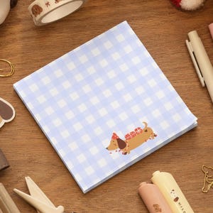 May include: A square notepad with a light blue and white gingham pattern. A small illustration of a brown dachshund wearing a red party hat and carrying red gift boxes is positioned in the lower right corner of the notepad pages.