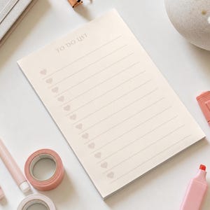 May include: A rectangular notepad with a cream-colored background featuring the text TO DO LIST at the top. The page includes twelve horizontal lines, each preceded by a small, light-colored heart icon for checking off tasks in a list format.