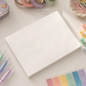 May include: A rectangular notepad with white paper featuring a light gray dot grid pattern. The notepad is positioned centrally among various pastel-colored stationery items, including pens, washi tape, paper clips, and sticky note tabs.