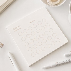 May include: A square off-white paper notepad featuring a printed goal tracker with the text GOAL, START DATE, and END DATE. The grid consists of twenty-five empty circles arranged in five rows and five columns, printed in a light brown ink color.
