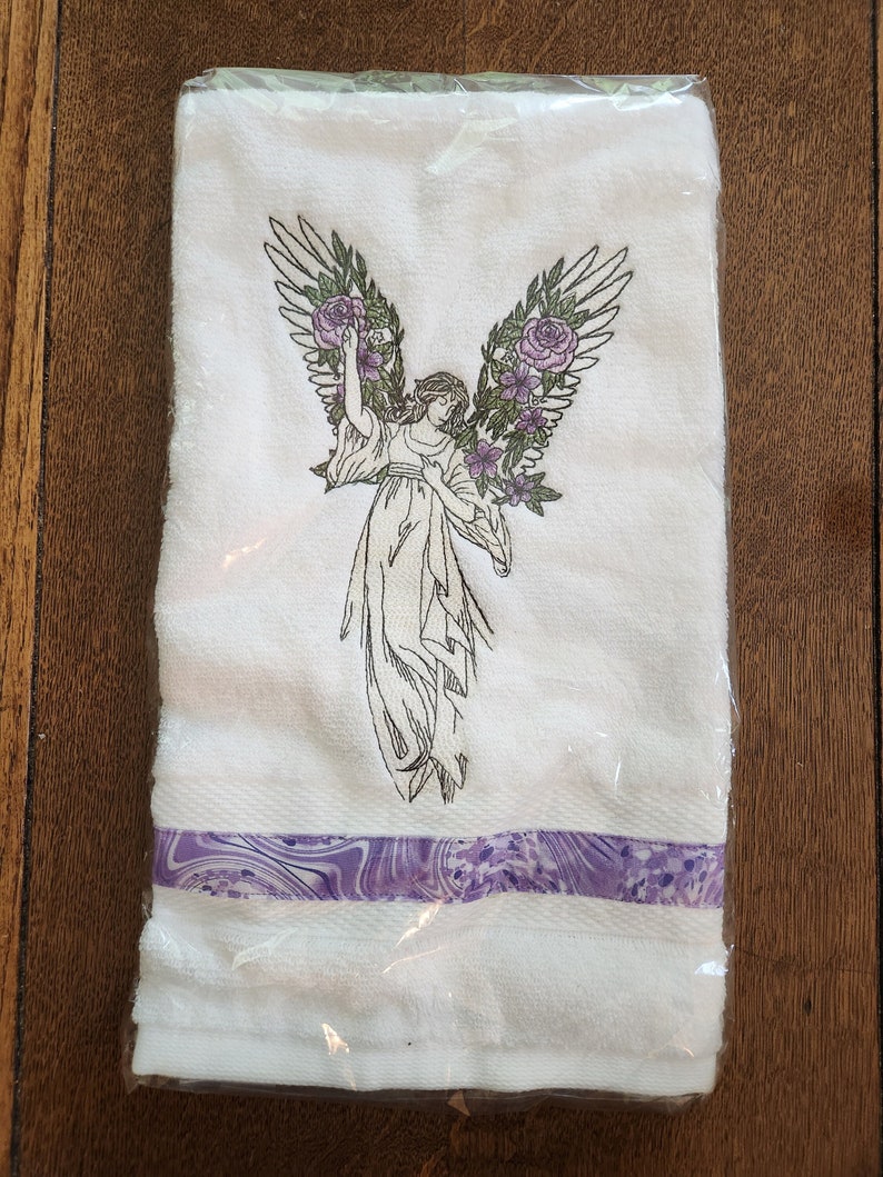 Op de afbeelding: Witte handdoek met een paarse en witte gestreepte rand. De handdoek heeft een geborduurd ontwerp van een engel met vleugels en bloemen.
