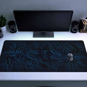 Puede incluir: Alfombrilla de ratón para ordenador negra y azul con diseño de placa de circuito. La alfombrilla rectangular presenta una red de líneas azules sobre un fondo negro. Un ratón de ordenador gris está sobre la alfombrilla, con un monitor y altavoces en el fondo.