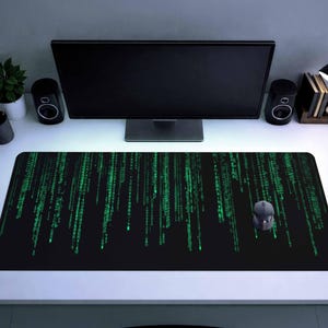 Puede incluir: Alfombrilla de escritorio rectangular negra con un patrón de lluvia digital verde, un ratón de ordenador y un monitor de ordenador. La alfombrilla mide aproximadamente 91 cm de largo y 30 cm de ancho. La configuración incluye altavoces, una lámpara de escritorio y una pequeña planta en maceta.