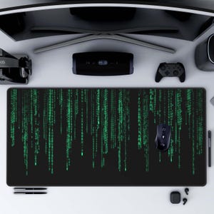 May include: A black gaming mouse mat with a green matrix code design. The mouse mat is on a white desk with a black computer monitor, a black speaker, a black video game controller, and a black and silver wireless headset.