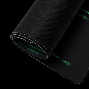Puede incluir: Alfombrilla de rat&oacute;n para juegos negra enrollada con costuras blancas. La alfombrilla presenta texto y s&iacute;mbolos verdes, con una superficie texturizada. El borde de la alfombrilla es visible, mostrando las capas y las costuras.