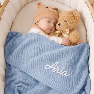 May include: A sleeping baby wearing a tan bear hat and a white onesie, nestled in a crib with a teddy bear. The baby is covered with a light blue knitted blanket embroidered with the name "Aria" in white script.