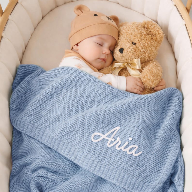 May include: A sleeping baby wearing a tan bear hat and a white onesie, nestled in a crib with a teddy bear. The baby is covered with a light blue knitted blanket embroidered with the name "Aria" in white script.
