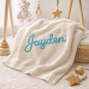 May include: Cream-colored knitted baby blanket with the name "Jayden" embroidered in turquoise script. The blanket is folded and resting on a white rug, with wooden baby toys in the background.